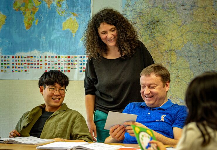 Ein Lehrerin mit Teilnehmern von DIE NEUE SCHULE lachend in einem Deutsch Intensivkurs vor Landkarten.